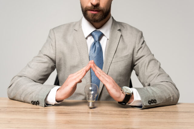 partial view of businessman showing protection gesture above light bulb isolated on grey