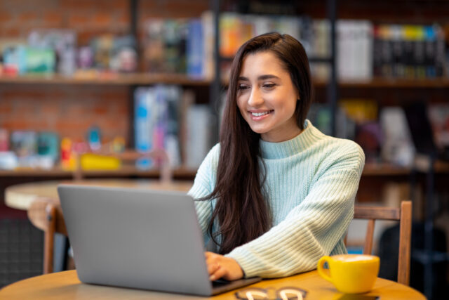 woman working on laptop computer in cafe, looking and smiling at screen, copy space. Happy female freelancer working distantly, chatting with clients or surfing internet