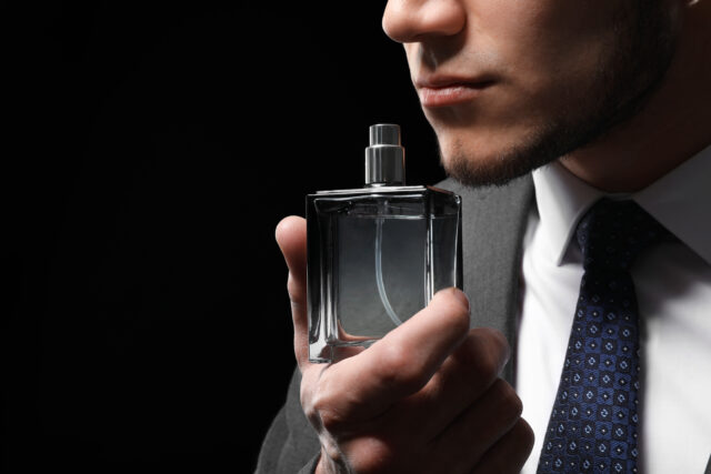 Handsome man in formal suit and with bottle of perfume on dark background, closeup